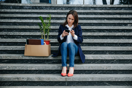 An Unhappy Woman, Recently Facing Dismissal, Sits on Staircase Searching for Job Opportunities on her Smartphone.の写真素材