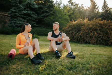 Two athletes, a man and woman, wearing fitness clothes, are sitting outside, talking and winding down together after their training. The man has a water bottle, and the woman carries a fitness mat.の写真素材