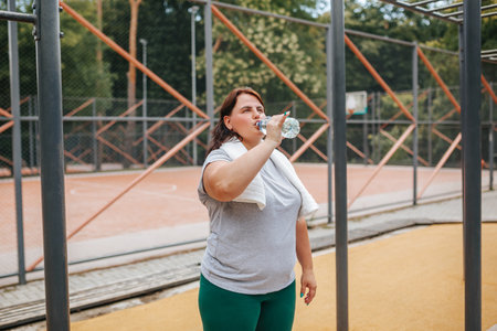 An overweight woman, dedicated to her fitness journey, takes a refreshing sip of water after an outdoor workout session, embodying the essence of a healthy lifestyle.の写真素材
