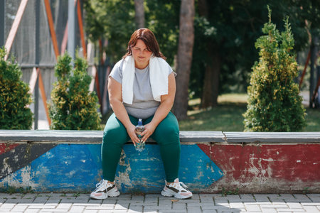 Portrait of an American plus-size female in sportswear, taking a break outside. Her water bottle symbolizes her commitment to a healthy lifestyle and weight loss.の写真素材