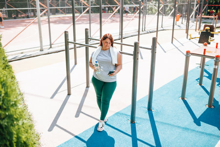 A plus size woman, after her outdoor exercise, enjoys her smartphone. Her smile of happiness exudes satisfaction, with a water bottle nearby.の写真素材