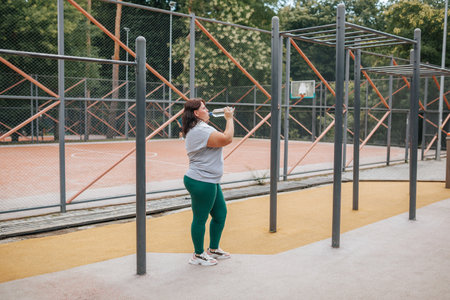 This real life example of a plus size woman exemplifies the beauty of determination and healthy living, as she quenches her thirst with water during an outdoor exercise session.の写真素材