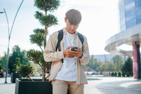 Asian Student, Full of Joy, Uses His Smartphone for an Internet Chat Session, in a Natural Setting. Happy Student with Mobile Phone and School Bagの写真素材