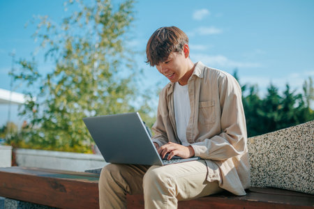 A committed Asian Korean guy, diligently working on his laptop, showcases the essence of a modern-day student. Amidst the greenery of a peaceful park, hes engaged in the process of learningの写真素材