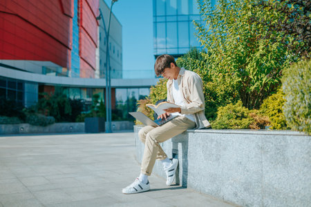 Knowledge Seeker A young Asian student seeks knowledge passionately, using both a laptop and a book. He finds inspiration outdoors, under the open sky, amidst the beauty of the campusの写真素材