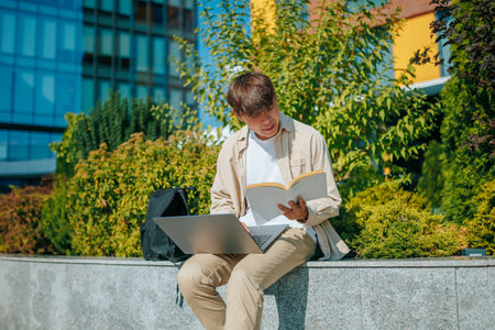 Focused on Learning A young Asian Korean man sits on a park bench, studying diligently on his laptop and engrossed in a book. His commitment to education shines through.の写真素材