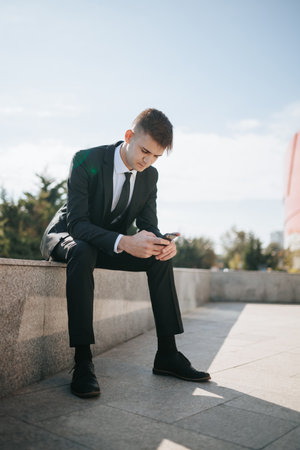 urban business district, a young Caucasian American businessman sits intently, dressed in a sharp business suit. Hes surrounded by towering buildings as he focuses on his smartphone,の写真素材