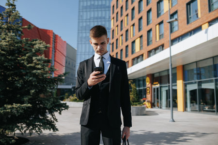 In the heart of the bustling urban business district, a young Caucasian American man dressed in a sharp business suit stands against a backdrop of towering buildings. engrossed in his smartphoneの写真素材