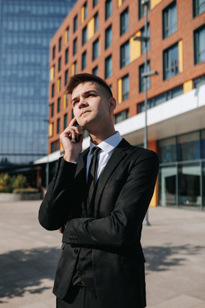 vertical shot In an outdoor urban environment, a young Caucasian male, dressed in a business suit, talks on his smartphone, highlighting the daily life of a young white collar worker.の写真素材