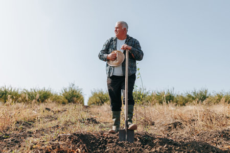 Standing Strong in the Fields A Mature Farmer Embraces the Serenity of His Farmland, Ready to Begin His Days Work with a Shovel. Standing Strong in the Fields The Senior Farmer and His Trusty Shovelの写真素材