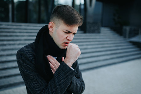 Outdoor Struggle A young man endures flu symptoms, constantly sneezing and coughing, reflecting discomfort while outside in the city.の写真素材