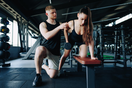 A supportive male personal trainer assists a woman in lifting a dumbbell, fostering a positive and empowering gym experience. Personalized Workout Male Trainer Assists Woman in Gym Trainingの写真素材