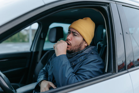 Witness a young chauffeur battling the flu behind the wheel in this 4k image. The bearded Hispanic American man, wearing a hat, sneezes and coughs on busy city streets, highlighting the impactの写真素材