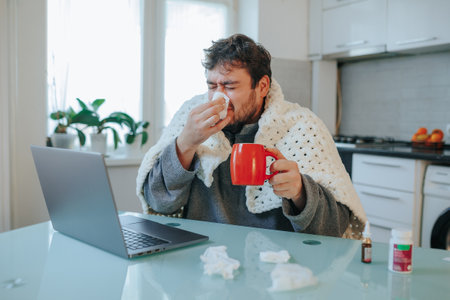In a vivid image, a young man with a beard contends with the flu, wrapped in a duvet on a kitchen chair. Shivering and wiping his nose, it authentically captures seasonal diseases.の写真素材