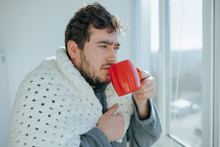 Objective realism bearded man wrapped in a blanket at home, dealing with flu on a kitchen chair, engaged in the act of drinking from a cup. Genuine Image Bearded Man Coping with Seasonal Diseasesの写真素材