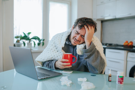 Chilled Workspace Blues Bearded man, tousled hair, flu stricken, seeks warmth under duvet, beside laptop, battling a cold day at home.の写真素材