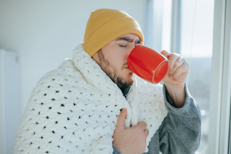 Nuanced reality stressed young man coping with flu, wrapped in a blanket at home, sitting on a kitchen chair, drinking from a cup. Nuanced Scene Young Man in Blanket Confronts Flu at Homeの写真素材