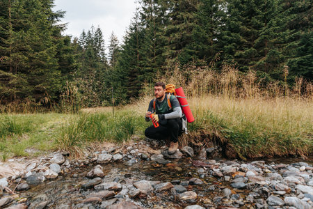 Sip of the Wilderness Young Backpacker Engages in a Tranquil Pause, Filling His Bottle from a CrystalClear River.の写真素材
