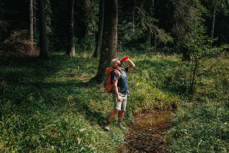 Join a Caucasian traveler on a trekking exploration, navigating through a wooded landscape, and witness a rejuvenating break as he quenches his thirst, drinking water from a bottle.の写真素材