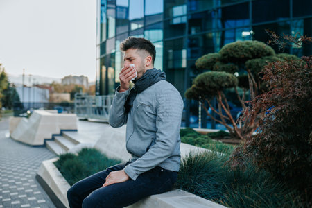 Capturing the flu ordeal of a young bearded Hispanic American man, sneezing and coughing uncontrollably, showcasing the discomfort of seasonal illness on the urban streets.の写真素材