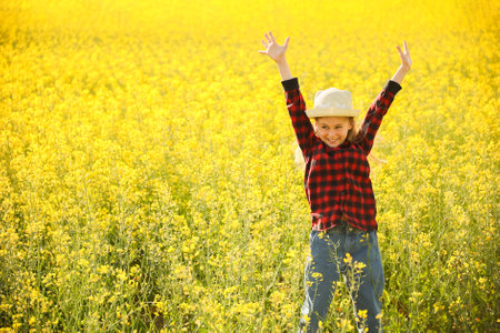 front view of little kid girl with hat looking at camera raise your hands up in flowering rapeseed land. happy little girl jumps upの写真素材