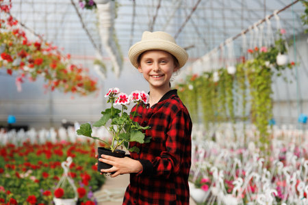 preteen kid happy girl with hat and a flower pot in her hand. smiling child looking at camera is in the greenhouse with many flowers, Copy spaceの写真素材