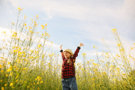 happy little girl with hat with her hands up. farmland with yellow flowering rapeseed, kid with outstretched armsの写真素材