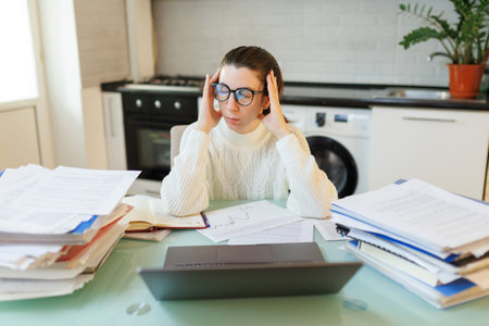Taking a momentary break, a woman, visibly tired and wearing glasses, reflects on her work at home, surrounded by her laptop and various documents.の写真素材
