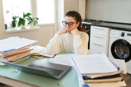 In her home workspace, a tired woman wearing glasses takes a break from her work on a laptop, surrounded by papers that highlight her industrious efforts.の写真素材