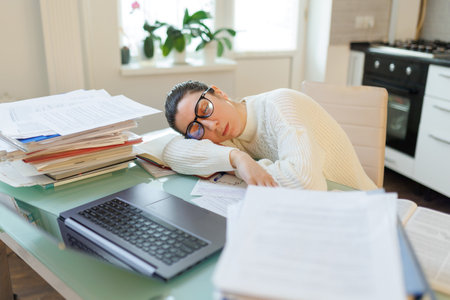 Exhausted from work, a woman wearing glasses takes a break at her home workspace, surrounded by her laptop and a table filled with documents.の写真素材