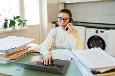 A tired woman, wearing glasses, pauses from her remote work, speaking on the phone amidst a table cluttered with documents and a laptop.の写真素材