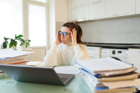 In her home environment, a woman with glasses takes a break from her work, the fatigue evident from hours spent on her laptop amidst scattered documents.の写真素材