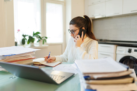 In her home workspace, a woman takes a break from laptop work, talking on the phone while surrounded by scattered documents and a computer.の写真素材