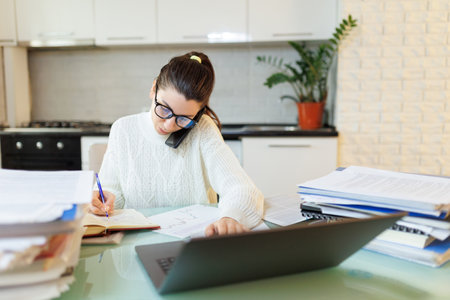 Taking a breather, a woman works at home on her laptop, talking on the phone, with documents spread across the table in her home office.の写真素材