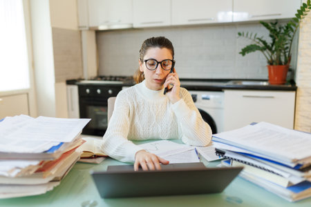 A woman in glasses, tired from work, sits at her home office table, engaged in a phone conversation amidst scattered documents and a laptop.の写真素材