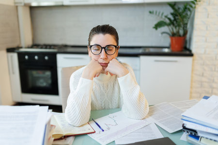 In her home workspace, a woman wearing glasses takes a momentary respite from work, her laptop and scattered documents capturing the essence of her efforts.の写真素材