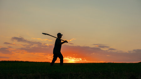 In the tranquil beauty of a rural evening, the silhouette of a genuine farmer emerges, walking with a shovel in the vast field. The dramatic sky sets the scene aglow with vibrant colorsの写真素材
