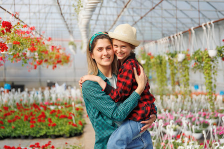 Cheerful kid girl in red square shirt and hat sitting on mothers hands. happy pretty daughter and mom family looking at camera and posing in greenhouse. gardeners family, copy spaceの写真素材