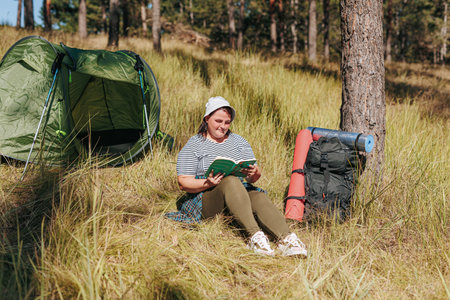 Join a plus size American hiker in the woods, capturing moments of tranquility as she reads a book during a hiking break. Witness the female traveler finding comfort next to her backpack and tent,の写真素材