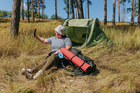 Snapshot of Serenity Plus Size Woman Selfie Moment on a Relaxing Hiking Break. Savoring Serenity overweight female Relaxing Hiking Break, Capturing Moments with a Selfie by the Tent and Backpackの写真素材