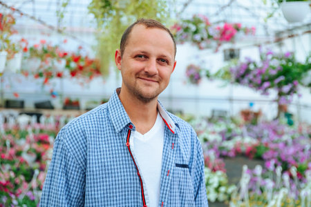 A young handsome farmer stands in a greenhouse and looking at camera. The head is slightly tilted to the left. The man smiles a calm smile. The agronomist is dressed in a plaid shirt. Copy spaceの写真素材