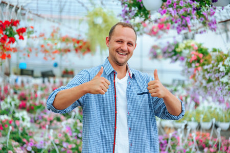 A happy young guy stands in greenhouse with flowers and smiles broadly, looking at the camera, shows thump up. The man man wearing a plaid shirt and has good mood. Around the flowers in pots. Copy spaceの写真素材