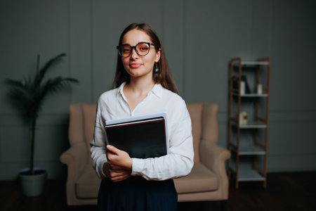 Young woman adult girl brunette glasses stand in office and holds folder with documents in her hands, look in camera smile. Front view, stands in white shirt. end of working day, beginning.の写真素材