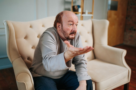 Front view aged man gesticulating with his hands trying to learn something with his eyes. Questioning look of elderly man. Confused look. Sitting indoors, at home. There are no thoughts. Lonely man.の写真素材