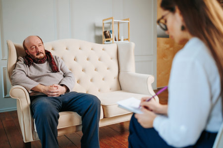Elderly man sits on a comfortable couch in a bright psychologist office. Client has senile disorders, a feeling of loneliness, a bad mood, constant fatigue and exhaustion of the nervous system.の写真素材