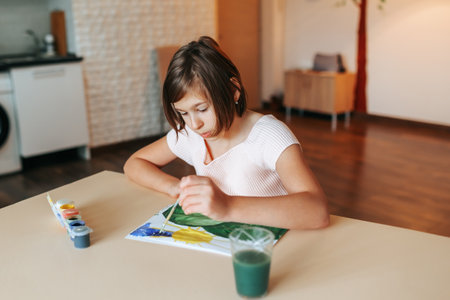 Home at the living room child sits at the table and draws a picture with paints and a brush in the album. Both hands on the table, attentive look. Next to a glass of water and paint. Front view.の写真素材