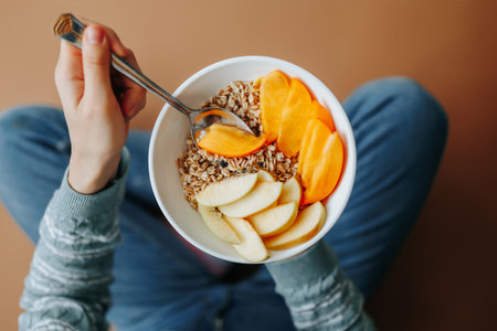Top view woman sits on the floor at home and holds in her hands a plate with healthy and tasty breakfast, granola muesli with fruits. Cereals, fruits, vegetables are good for digestion and good mood.の写真素材