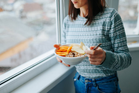 Front view unrecognizable woman standing at the window at home holding a plate and a spoon with a mixture of cereals with raw fruits. A young girl eats right and maintains a good mood and health.の写真素材