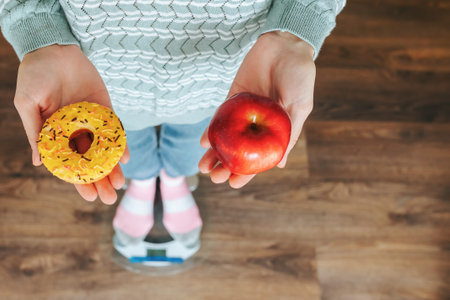 Close up donut and apple in unrecognizable hand of woman or girl standing on scales. Flour of choice. Diet. Sweet donut in hand, apple in hand, red apple, endless diet. Concept of beauty and health.の写真素材