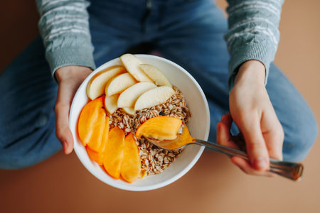 Top view plate with fresh fruits and muesli, unrecognizable woman eating breakfast Turkish sitting position. A girl sits on the floor at home and enjoys a delicious, healthy, nutritious breakfast.の写真素材